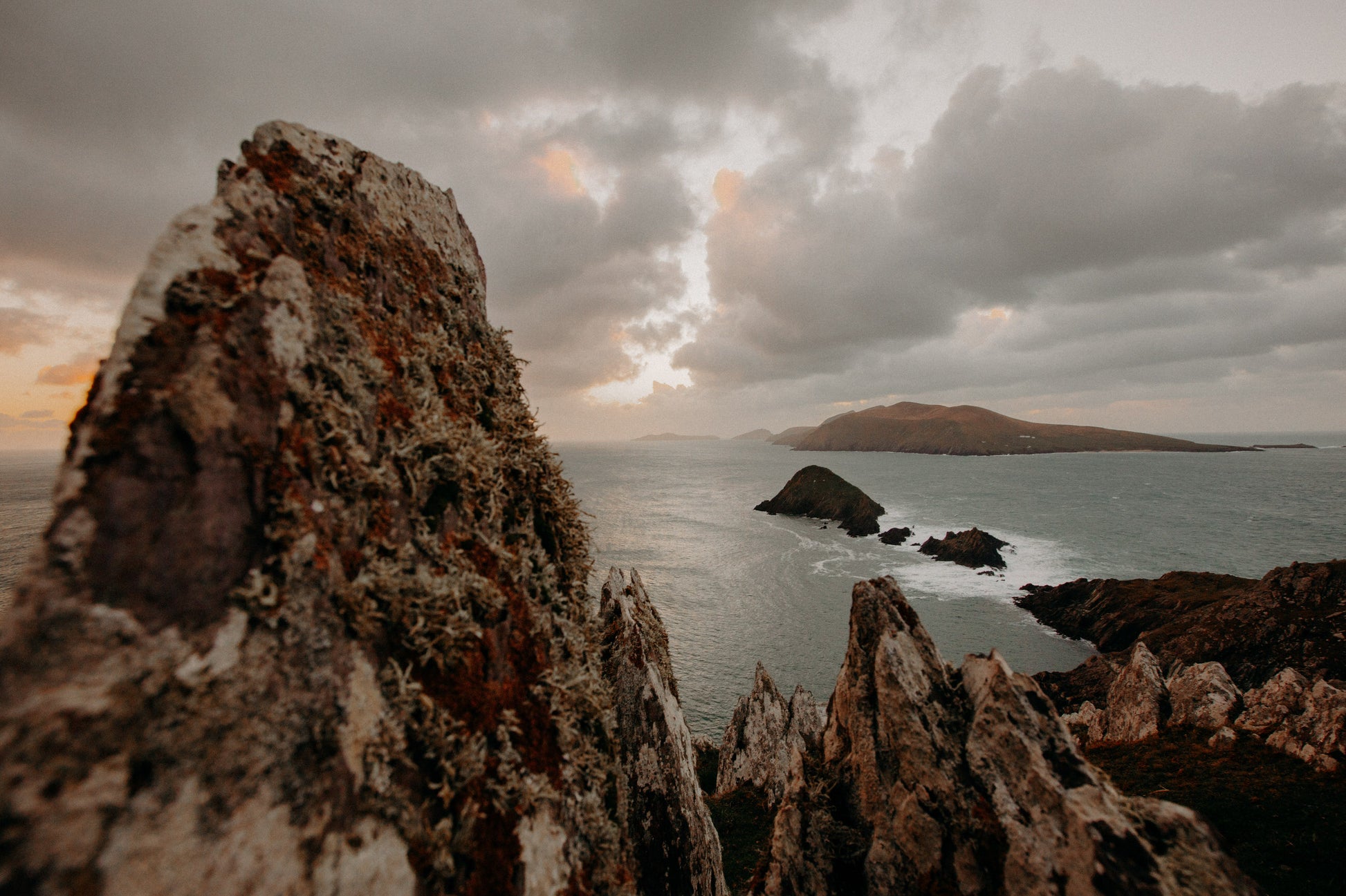 "Dramatic coastal landscape at Waymont (Clogher Head) on Slea Head, Dingle, Ireland. The rugged, lichen-covered rock formations in the foreground contrast with the vast Atlantic Ocean and distant islands. The scene is bathed in moody, golden light as the sun sets behind heavy clouds, casting an ethereal glow over the water and surrounding hills.