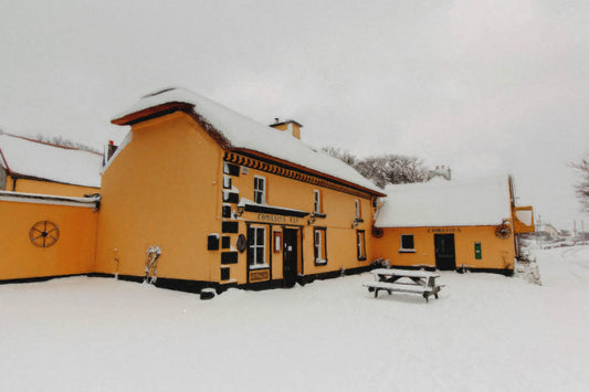 A picturesque winter scene of Tomásins Bar in Farrantooleen, Stradbally, County Kerry, Ireland, blanketed in fresh snow. The traditional thatched-roof pub, painted in warm ochre tones with black trim, stands out against the crisp white landscape. Snow covers the roof, ground, and a wooden picnic table outside, adding to the cozy, timeless charm of this historic Irish pub. The quiet surroundings and muted winter sky create a peaceful and nostalgic atmosphere, capturing the beauty of rural Ireland in winter.